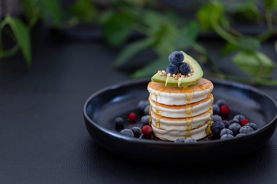 Pancakes with avocado and berries on top on a blacl plat with plants in the background