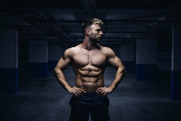 Fototapeta na wymiar Young handsome shirtless Caucasian muscular sportsman standing with hands on hips in garage at night and looking away. Urban life concept.