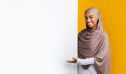 Smiling afro muslim woman in headscarf pointing at white advertisement board