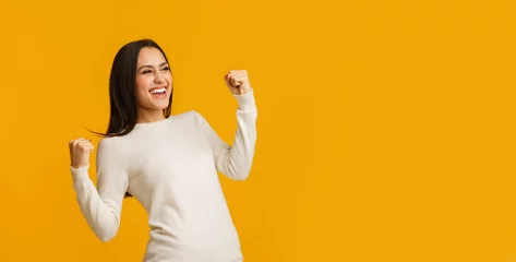 Foto op Plexiglas Euphoric brunette woman celebrating success with raised fists over yellow background © Prostock-studio