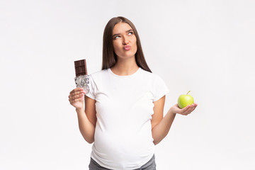 Fototapeta na wymiar Puzzled Pregnant Woman Choosing Between Chocolate And Apple In Studio