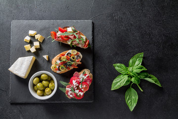 Traditional Bruschetta. Italian antipasti set with jamon, guanchial sausage, olives, cottage cheese, arugula and tomatoes on a black background
