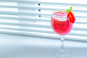 Fresh iced strawberry smoothie in wine glass decorated with slice of strawberry and mint against the window (shutters). Summer Slushy drink. Close up, copy space for text.