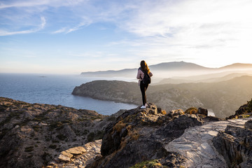 Beautiful woman standing on a cliff during sunset with the mediterranean sea in the background at Cap de Creus - obrazy, fototapety, plakaty