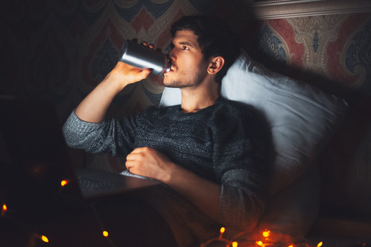 Night Portrait Of Young Man Lying On Pillow On Bed With Laptop, In Dark Room At Home With Garlands, Drinking Water From Reusable Aluminum Thermo Bottle.
