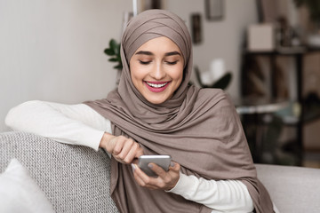 Cheerful muslim woman in hijab using smartphone at home