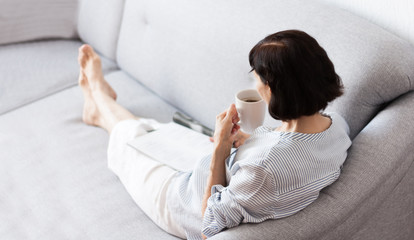 Middle-aged brunette woman with glasses on the gray sofa reading magazine with cup of coffee, soft focus comfort concept of loneliness.