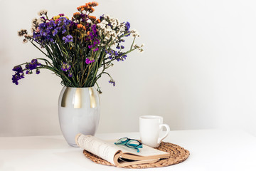 bouquet of flowers, open book and glasses, lemon tea in a white ceramic cup on a wicker stand on the table, cozy home interior.