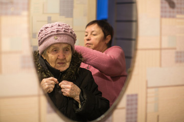 Reflection in the mirror, a woman helps put on her old mother's coat.