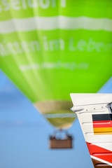 A flying hot air balloon in green, blurred in the background. In the foreground a German flag painted on the aircraft vertical fin.