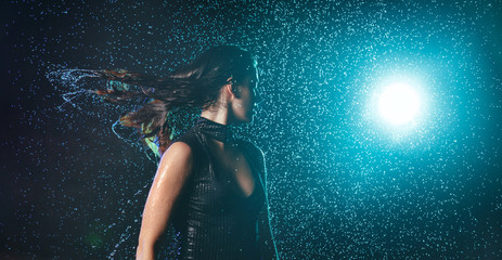 Fototapeta na wymiar Beautiful wet girl in a swimsuit shakes her head and streams of water flow down from her hair under raindrops with turquoise highlight on a black background.