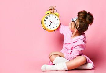 Yawning teenage girl holding an alarm clock while sitting in a housecoat on the floor.
