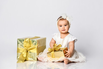 Baby girl in dress and headband, barefoot. She holding golden gift box, sitting on floor isolated on white. Close up, copy space