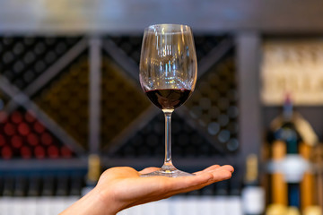 A glass of red wine on hand in selective focus close up view against wine bottles on X cube storage racks, tasting room blurred interior background