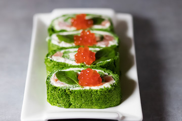 Spinach biscuit roll with cottage cheese, salted salmon and red caviar on a white rectangular plate on a gray table.