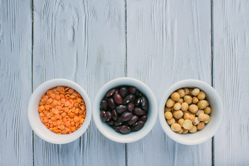 Three porcelain small cups with cereals top view: red beans, orange lentils and yellow peas on a white wooden background with space for copying