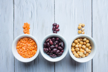 Three porcelain small cups with cereals top view: red beans, orange lentils and yellow peas on a white wooden background with space for copying