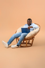 African-American man reading book while sitting in armchair against color background