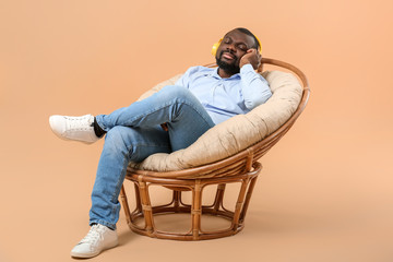 African-American man listening to music while sitting in armchair against color background