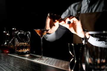 Professional bartender spraying on a brown alcoholic drink in a martini glass with orage zest juice