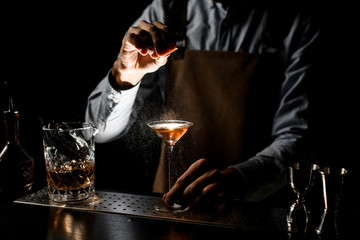 Professional male bartender spraying on a golden alcoholic drink in a martini glass with orage zest juice