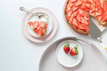A delicious, fresh tart with pudding cream and strawberries. A piece of grated on a pink plate. Everything on a white table.