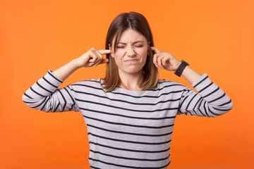Obraz na płótnie Canvas Don't want to hear it! Portrait of irritated woman with brown hair in long sleeve shirt standing grimacing and closing ears from noise, avoid conflict. indoor studio shot isolated on orange background