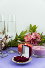 Marriage proposal and love concept for Valentine`s day, box with wedding or engagement ring with brilliant. Glass of champagne, romantic dinner, peony flowers and candles on trendy purple background