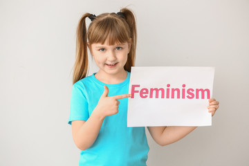 Little girl holding paper with word FEMINISM on light  background