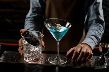 Professional bartender serving a blue alcoholic cocktail in a martini glass decorated with a dry spikelet