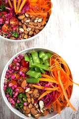 Top view of bowls of bran, pea seeds, carrots, green beans, cherry tomatoes, almonds and raisins on a light background