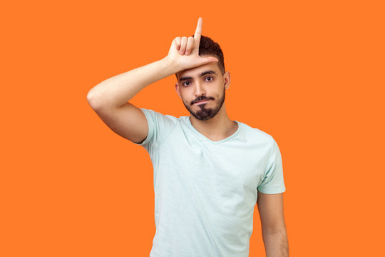 I am loser! Portrait of unlucky brunette man with beard in casual white t-shirt standing with grumpy face and showing loser gesture, upset over defeat. indoor studio shot isolated on orange background