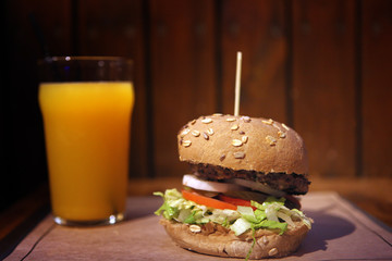 big homemade burger with grilled meat and orange juice. Big tasty burger lunch with beef BBQ. Lunch with Glass of orange juice on a wooden background. The concept of fast food and unhealthy fast food.