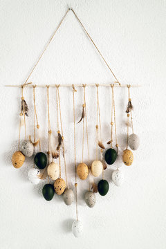 Hanging composition dream catcher of quail eggs and feathers hanging on a white wall at home. Easter home cozy concept