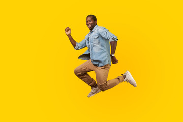 Fototapeta na wymiar Full length portrait of happy joyous man in denim casual shirt jumping or flying, hurry running to his dream, looking at camera with toothy smile. indoor studio shot isolated on yellow background