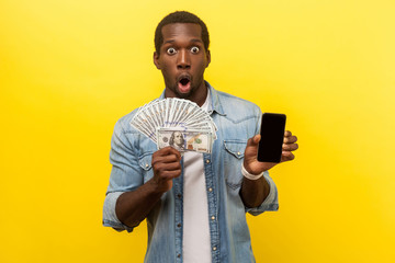 Wow, profitable service, online banking. Portrait of surprised excited man in denim shirt holding dollars and smartphone, making money in mobile application. studio shot isolated on yellow background