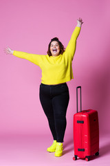 Cheerful plus size model with red suitcase, fat woman in black jeans and yellow jumper on pink...