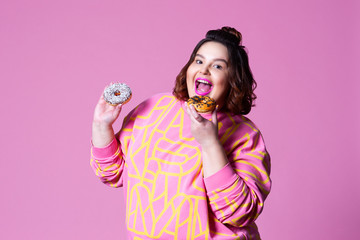 Cheerful plus size model eating doughnuts, fat woman overeats on pink background