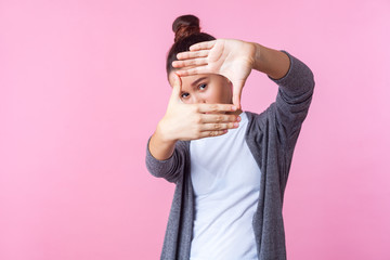 Portrait of curious brunette teenage girl with bun hairstyle in casual clothes looking through photo frame gesture made of hands, capturing moments. indoor studio shot isolated on pink background