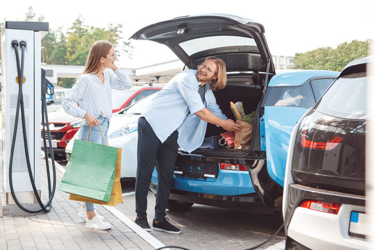 Traveling. Young couple traveling by electric car stop at charging station putting shopping bags into trunk looking at each other smiling cheerful