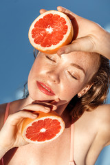 Summer Freestyle. Young woman with freckles and wet hair standing isolated on blue posing with grapefruit halves closed eyes smiling relaxeed close-up