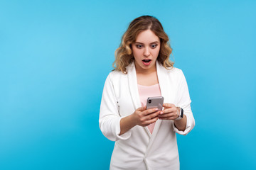 Fototapeta premium Portrait of surprised young woman with wavy hair in white jacket reading shocking message on cellphone, unbelievable news about sales, online shopping. indoor studio shot isolated on blue background