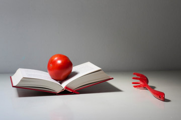 libro de tapas rojas con tomate rojo encima y cuchara de pasta roja al lado formando bodegón minimalista