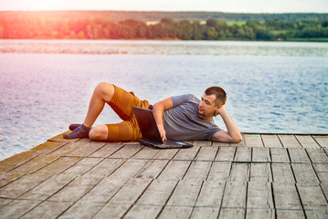 Young man sitting on a pier, using his laptop. Work and vacation.