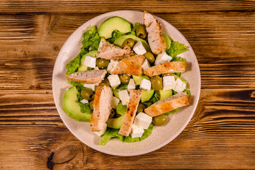 Fresh salad with chicken meat, feta cheese, avocado, green olives and lettuce leaves in ceramic plate on wooden table. Top view