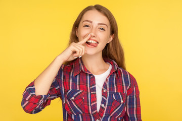 Portrait of funny childish ginger girl in checkered shirt picking her nose and demonstrating tongue with comical silly expression, pulling out boogers. indoor studio shot isolated on yellow background