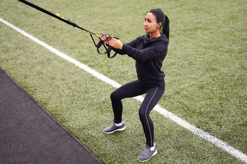 Girl athlete training using trx on sportground. Mixed race young adult woman do workout with...