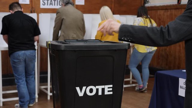 Voter inserts ballot paper into box at a Polling place - They are voting at an election in a public hall.&nbsp;The box says VOTE
