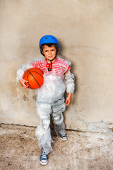 Child with basketball ball wear overprotecting super safe bubble wrap cover and helmet stand near the grey wall on the street