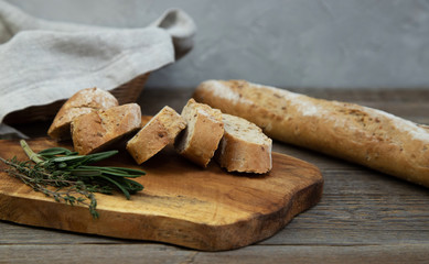 A whole-grain chopped baguette with herbs lies on a wooden board, next to a fresh sprig of thyme and rosemary herb. In the background a baguette and a wicker basket with bread on a wooden table